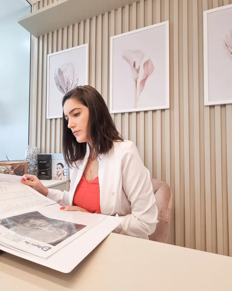 Dra. Ana Vieira analisando uma radiografia à mesa, com quadros de flores rosadas na parede