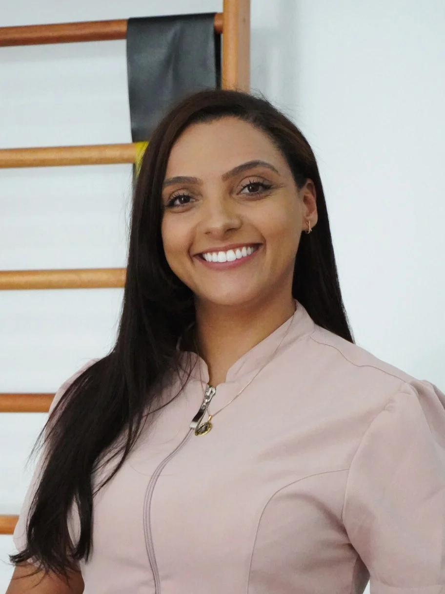 Dra. Vanessa Cruz, fisioterapeuta, sorrindo de uniforme rosé em frente ao espaldar da clínica