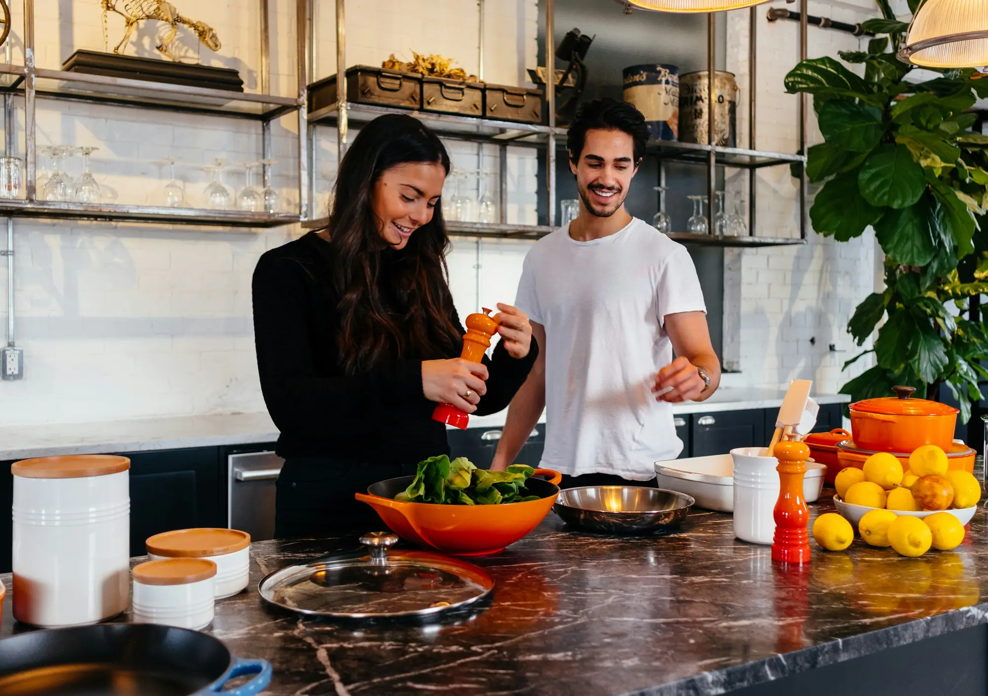 Casal cozinha junto em uma cozinha clara, com frutas e plantas